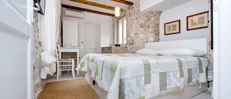 Un dormitorio con una cama, un escritorio y una silla. Hay una ventana, una puerta y una pared de piedra.