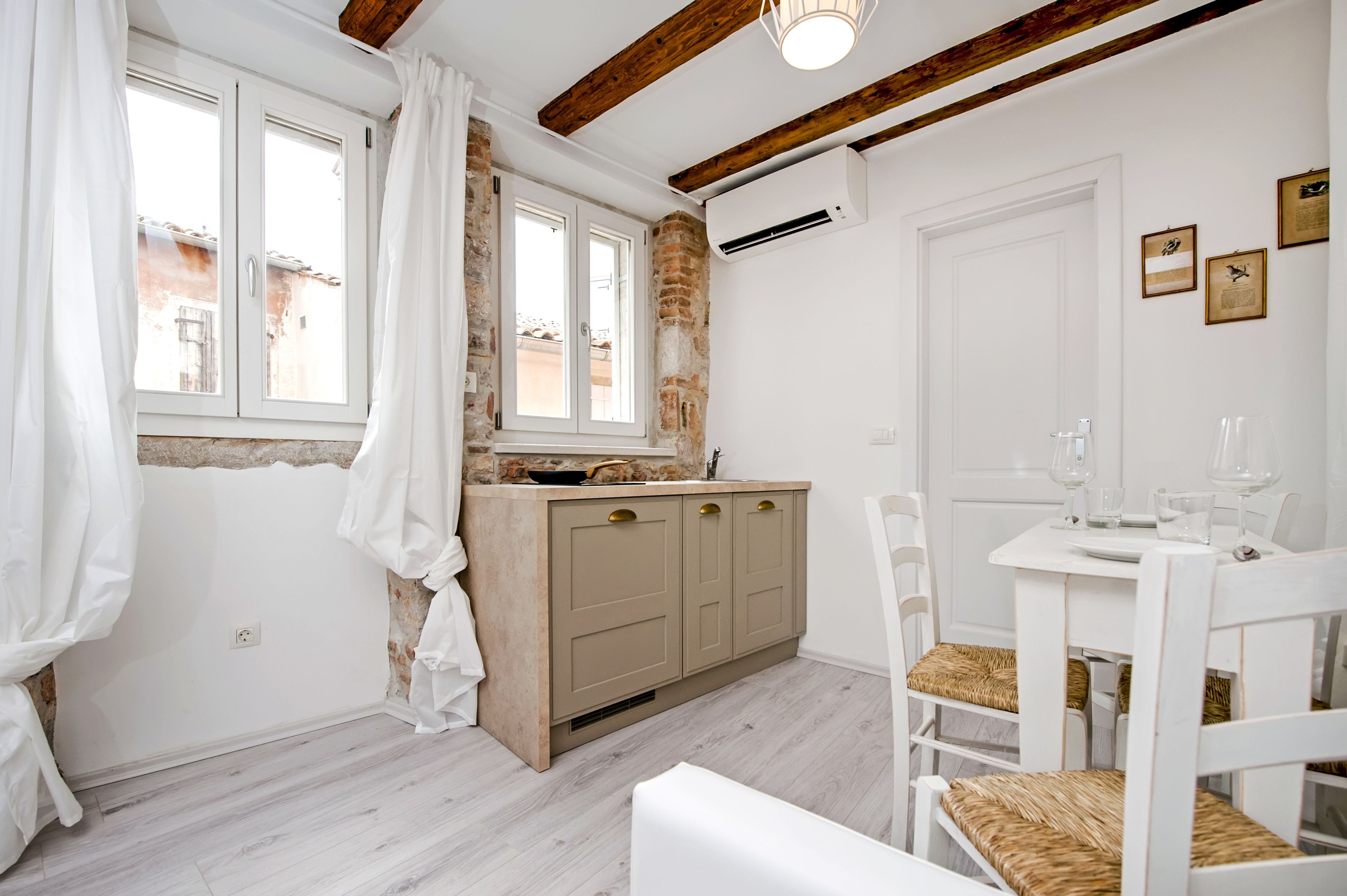 Una cocina con un armario de madera, una ventana con cortinas, una mesa blanca con sillas y un equipo de aire acondicionado en la pared.
