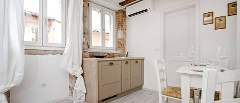 Una cocina con un armario de madera, una ventana con cortinas, una mesa blanca con sillas y un equipo de aire acondicionado en la pared.