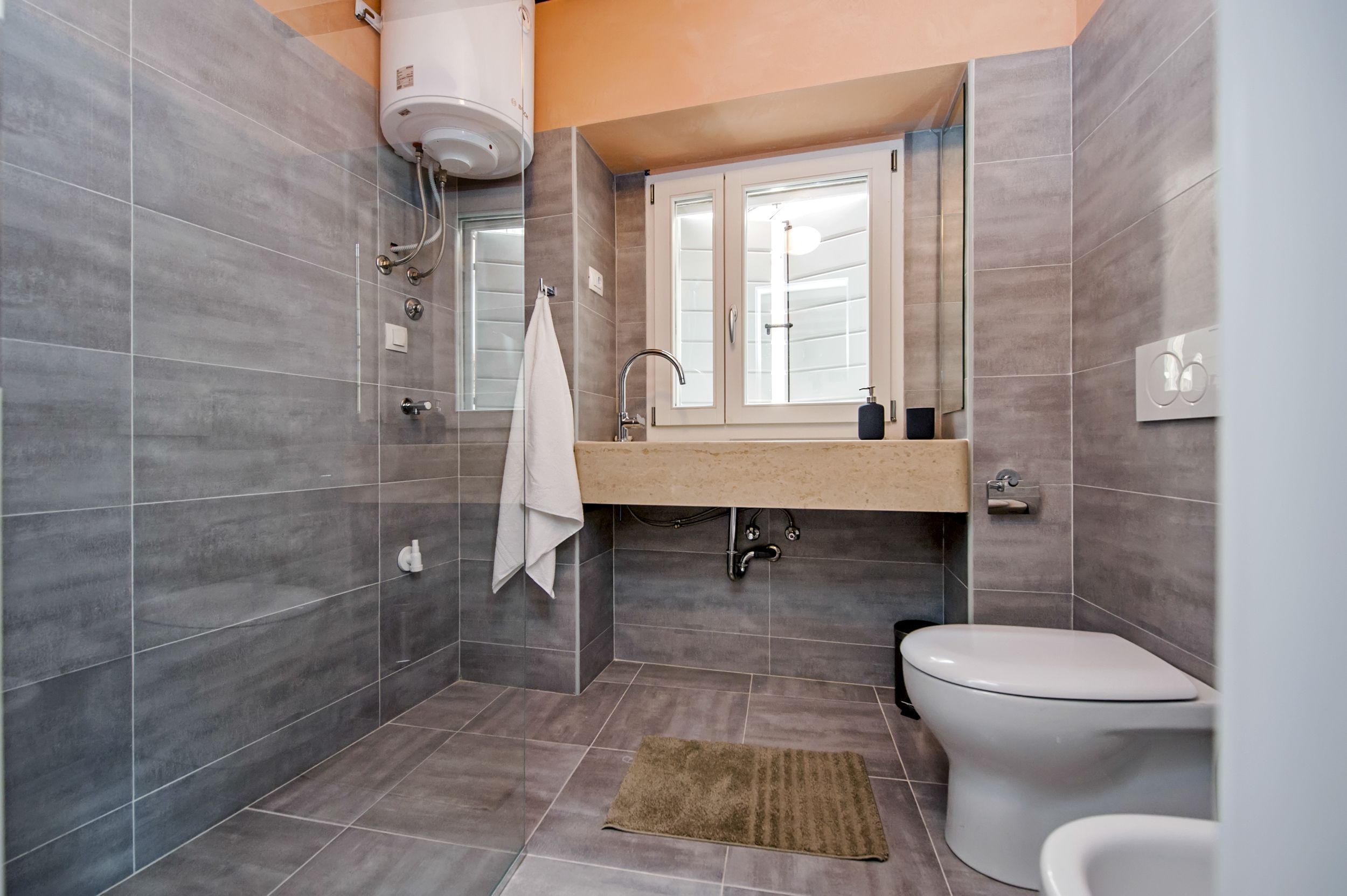 A modern bathroom with grey tiles, a white toilet, a sink with a mirror, and a towel hanging on the wall.