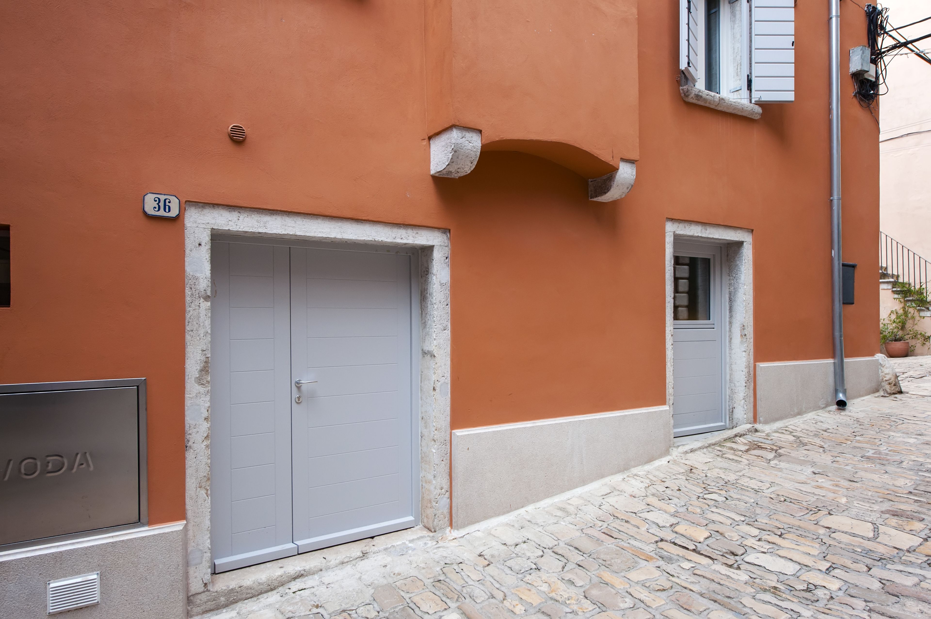 A building with a stone-paved street, a large grey door, and a smaller door with a window.