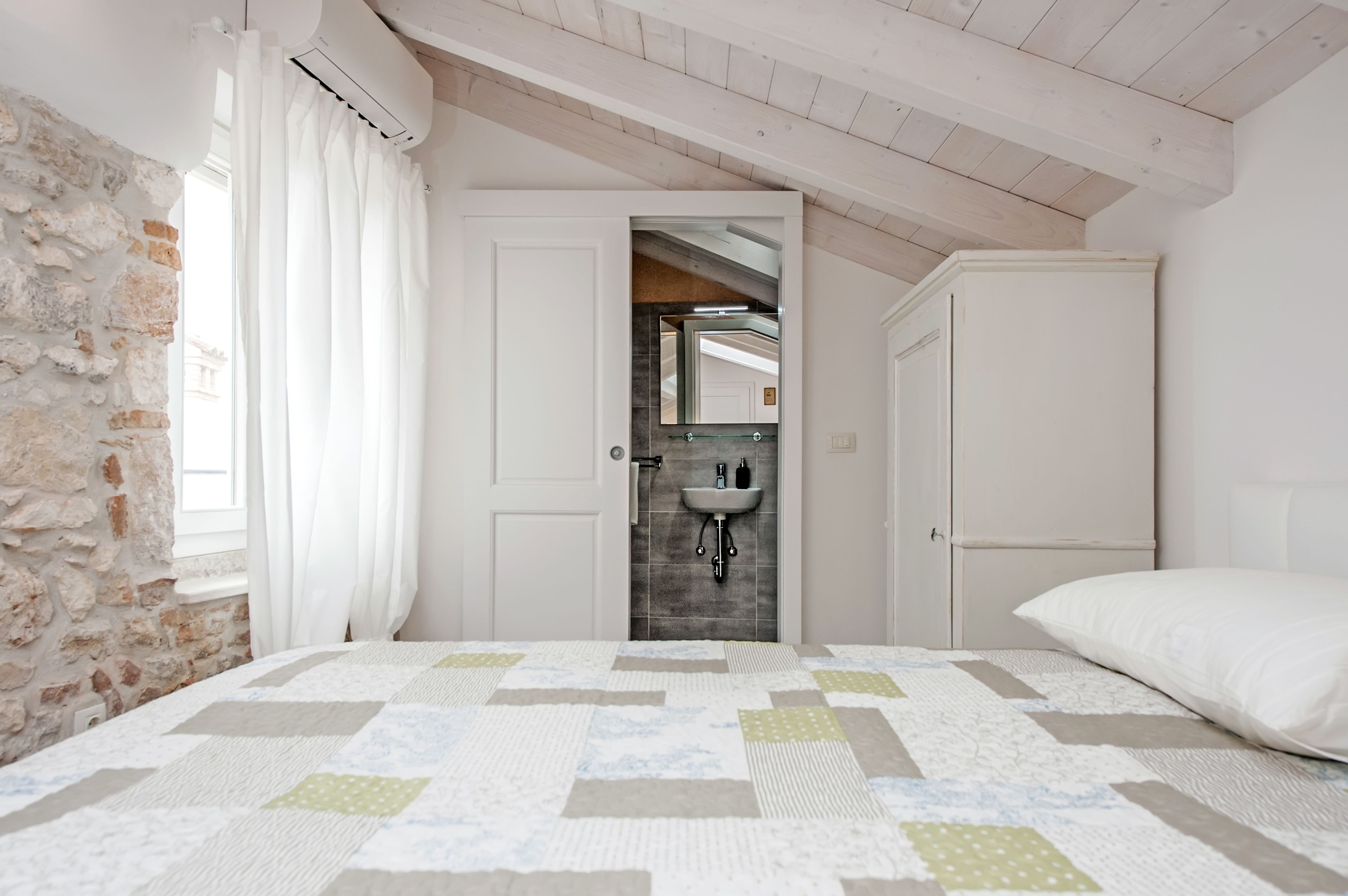 Ein Schlafzimmer mit gesteppter Bettdecke, einem weißen Kissen, einer Steinwand, einer weißen Tür, die ins Bad führt, und einem weißen Kleiderschrank.
