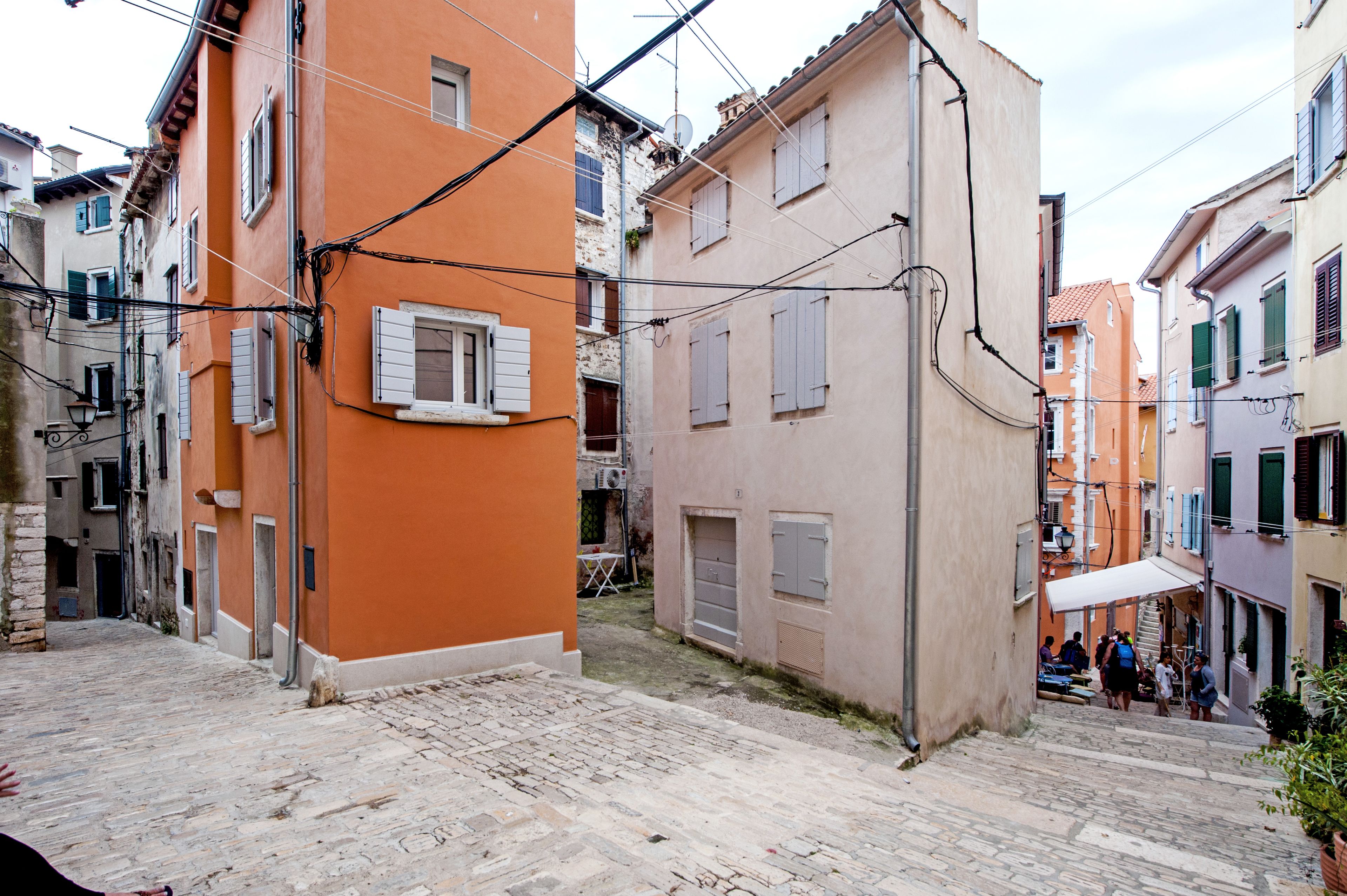 Una stretta strada acciottolata fiancheggiata da edifici colorati con persiane, un'antenna parabolica ben visibile e alcune persone in lontananza.