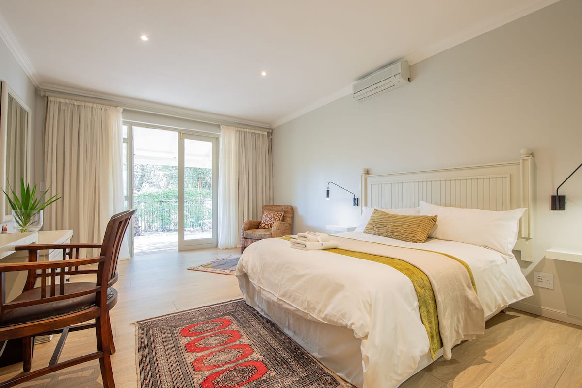 A bedroom with a large bed, a desk, and a chair. There is a window with curtains, a plant, and a view of greenery outside.