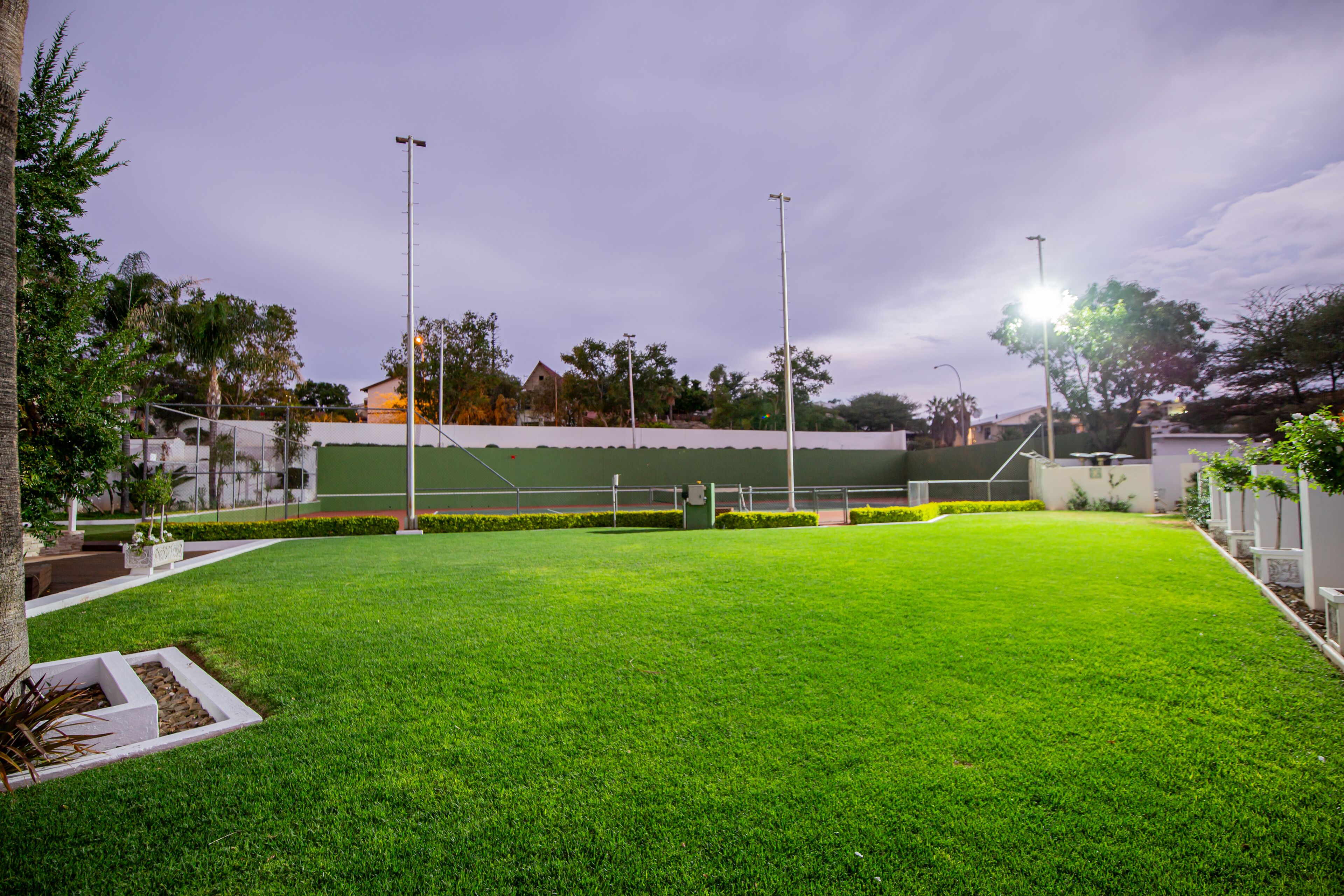 A well-maintained lawn with a tennis court, surrounded by trees and a building in the background.