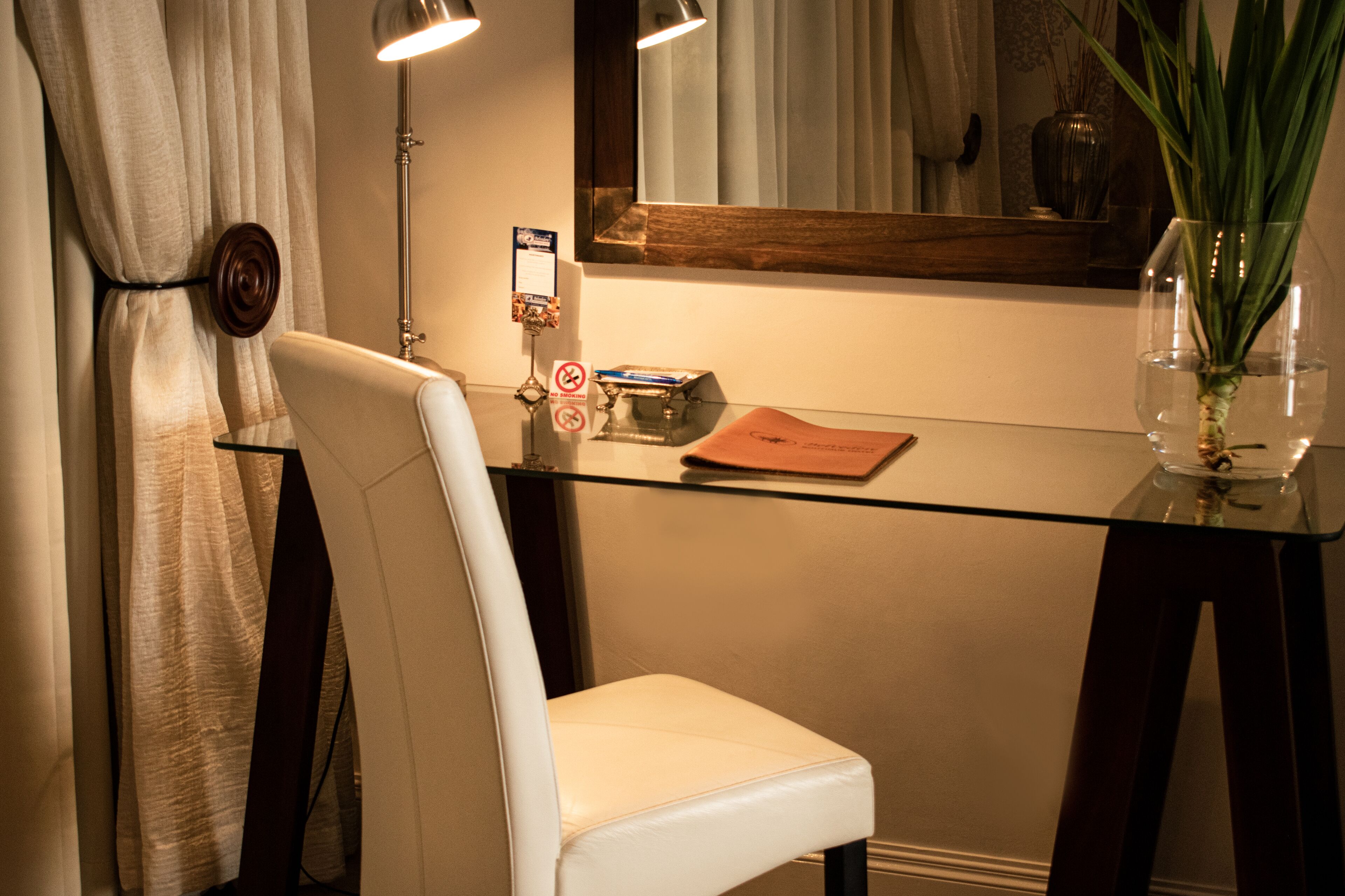 Une chambre d’hôtel avec une table en verre, une chaise blanche, un miroir et un vase avec une plante.