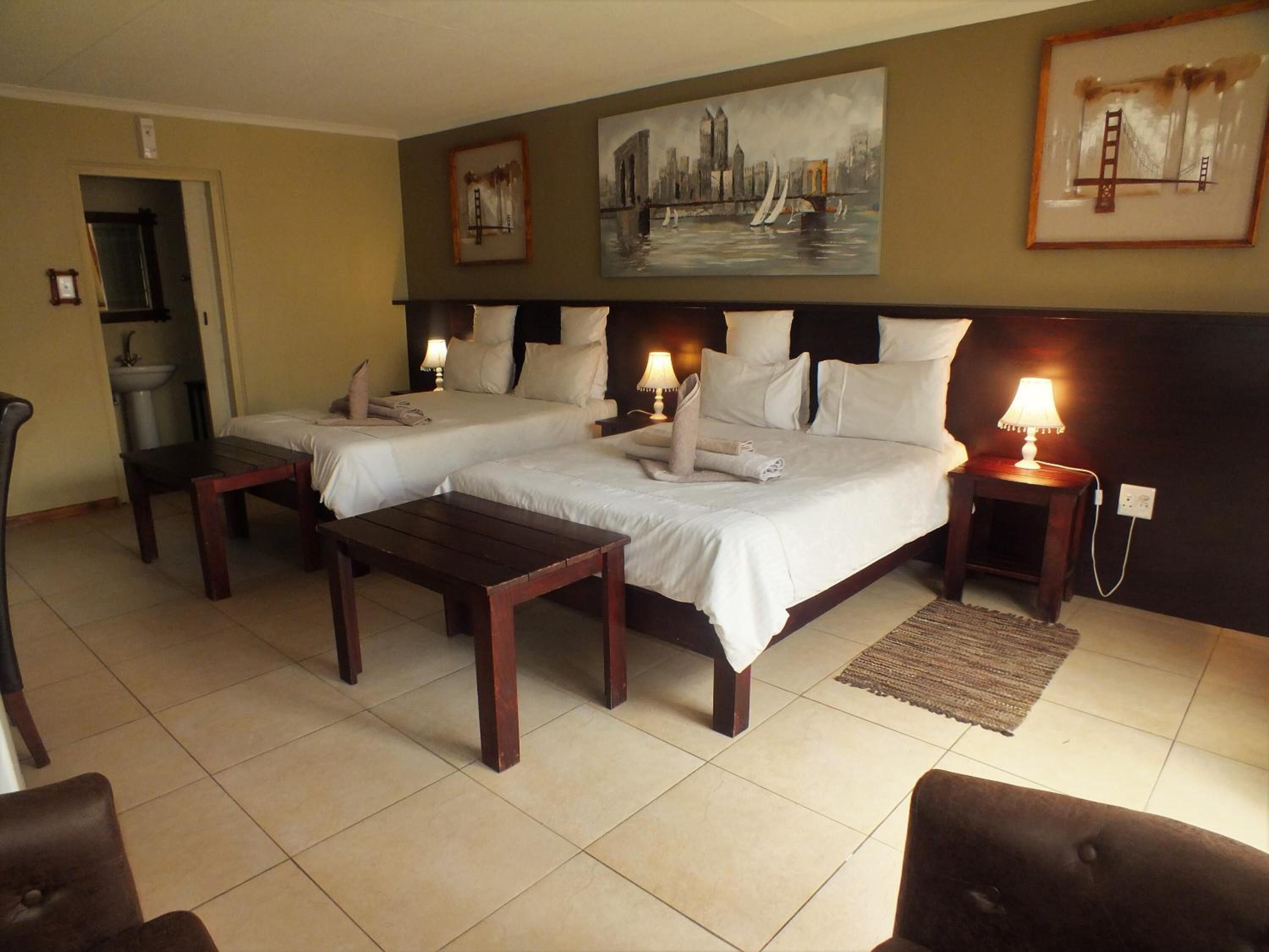 A hotel room with two beds, a desk, and a chair. There are paintings on the wall and a view of a cityscape.