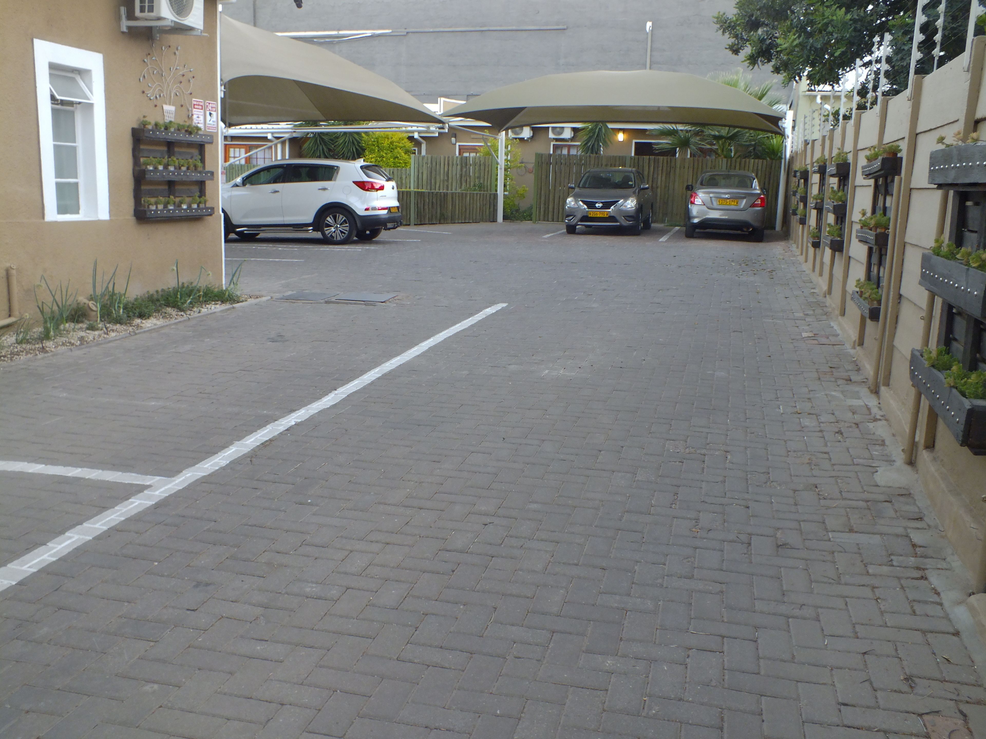 A parking area with a white SUV and two cars parked. There are planters with plants along the right side.
