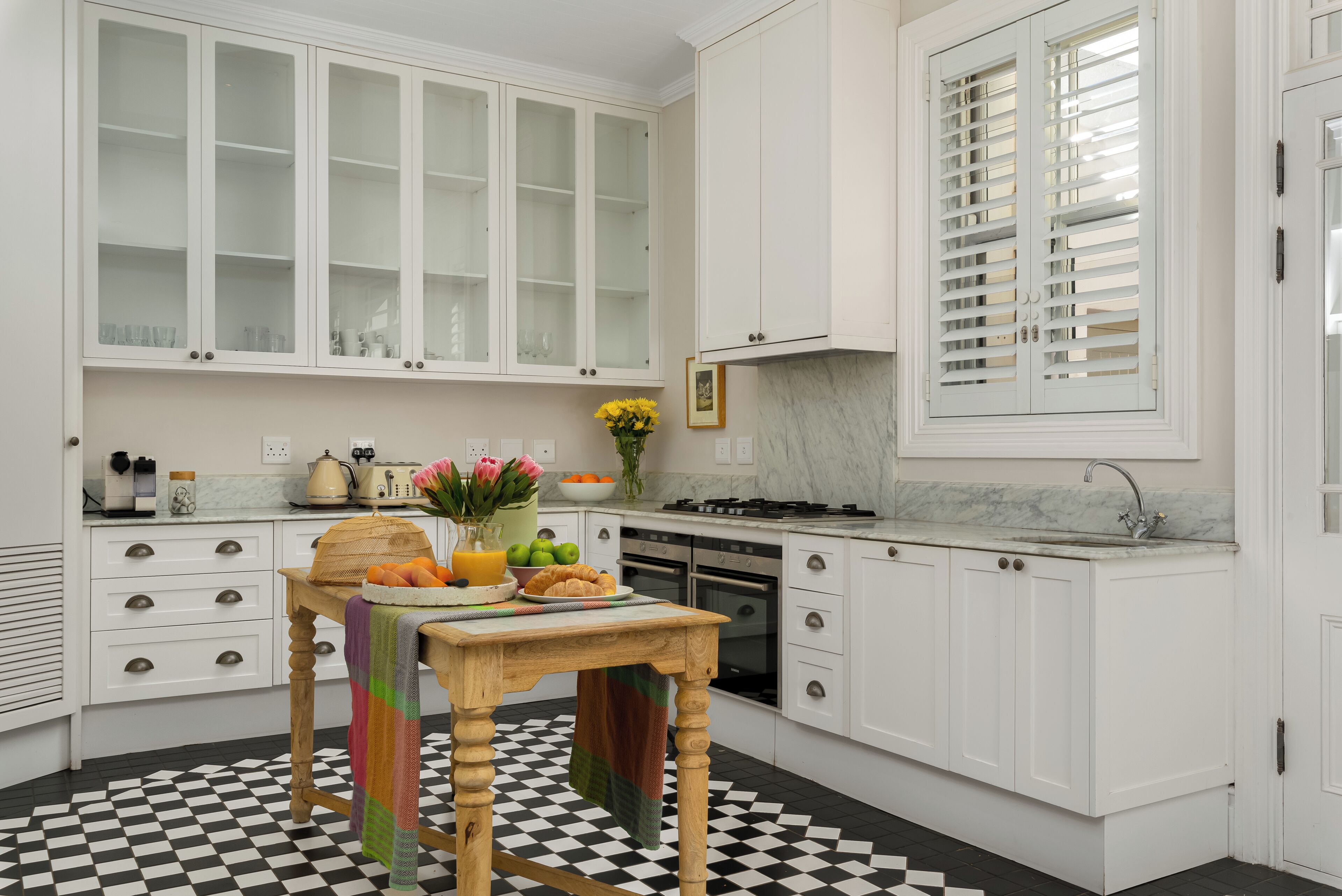 Una cocina con armarios blancos, piso a cuadros, una mesa de madera y una toalla colorida, y una ventana con persianas.