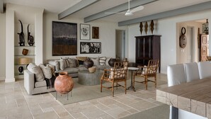 Uma sala de estar ampla com um sofá grande, uma mesa de centro e um espaço de refeições com cadeiras. Há um ventilador de teto, obras de arte nas paredes e um armário com objetos decorativos.