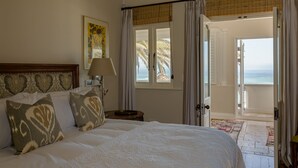 Quarto com uma cama grande, mesa de cabeceira, abajur, pintura e vista para o oceano através de uma porta aberta.
