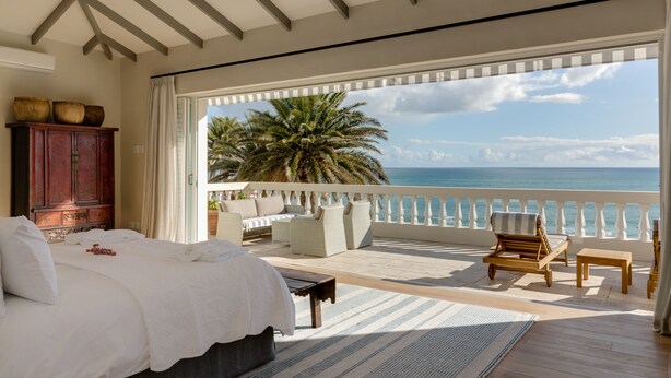 Quarto com cama grande, um banquinho de madeira, uma mesinha e vista para o oceano.