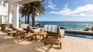 Área de piscina com espreguiçadeiras de madeira, uma mesa com garrafa e copos, e vista para o oceano.