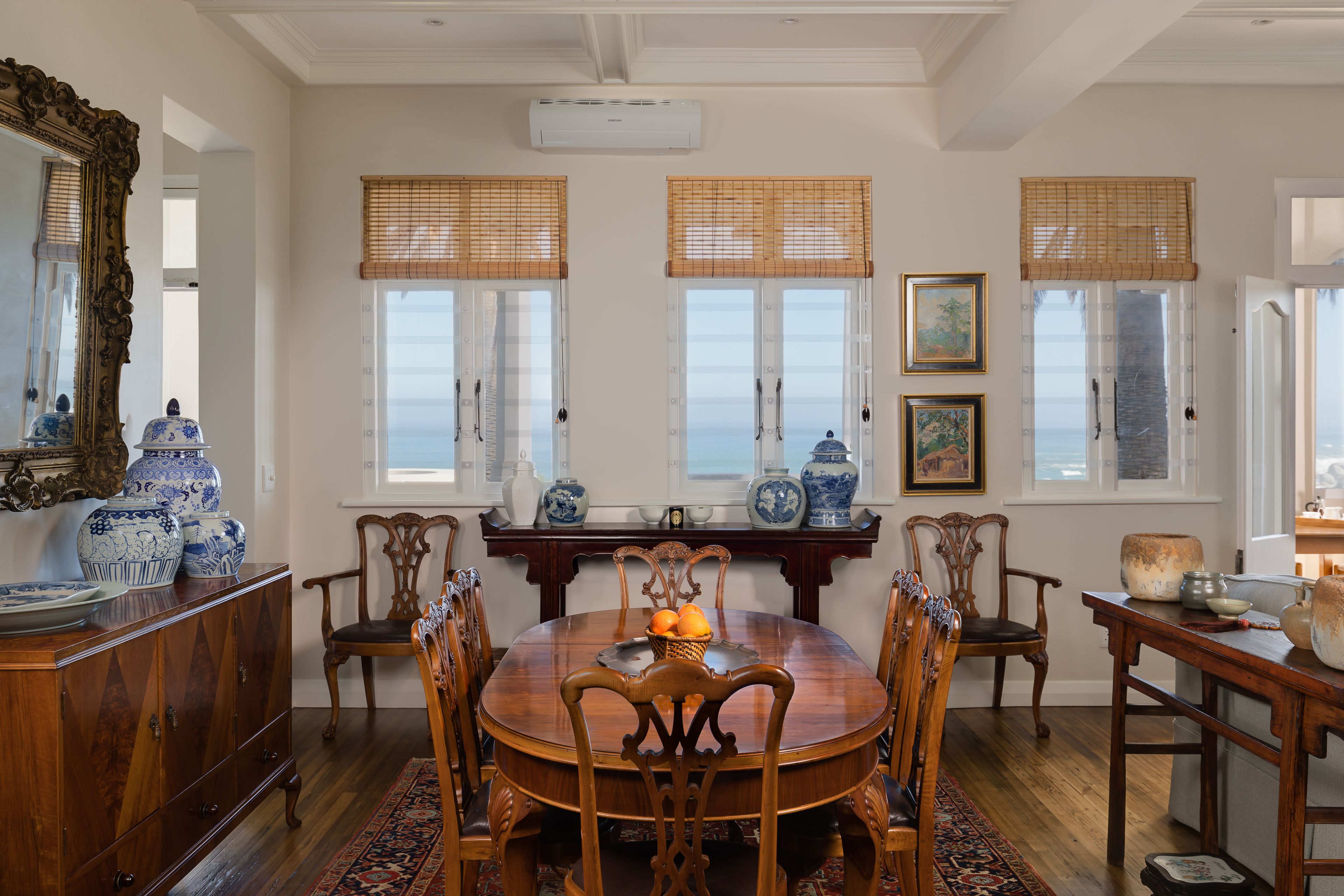 A dining room with a wooden table, chairs, and a view of the ocean through large windows with bamboo blinds.