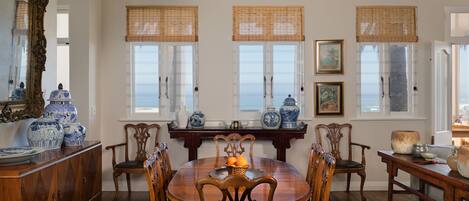 A dining room with a wooden table, chairs, and a view of the ocean through large windows with bamboo blinds.