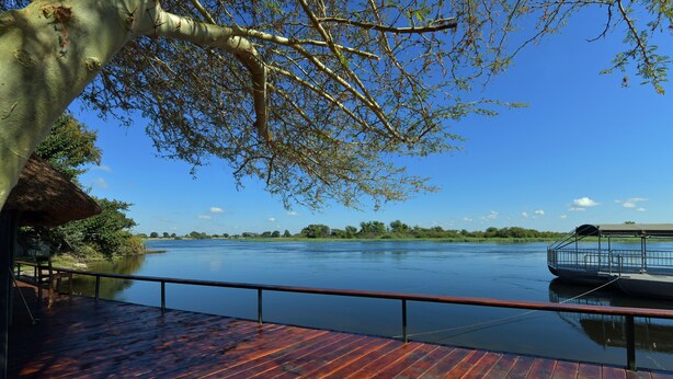 A wooden deck with a railing overlooking a calm river, surrounded by trees and a clear sky. - Zambezi Mubala Camp (Katima Mulilo)