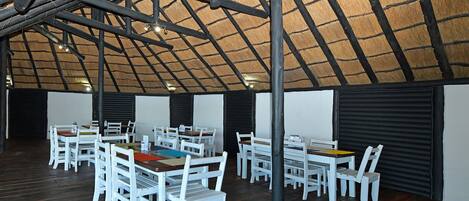 An indoor dining area with tables and chairs, a thatched roof, and a wooden floor.