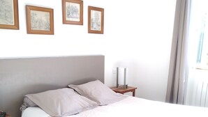 Un llit ben arropat amb un capçal, una tauleta de nit amb una làmpada i tres quadres al mur.