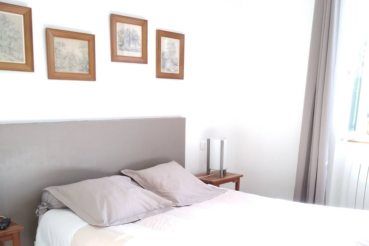 Un lit bien fait, avec une tête de lit, une table de chevet sur laquelle se trouve une lampe, et trois tableaux encadrés accrochés au mur.