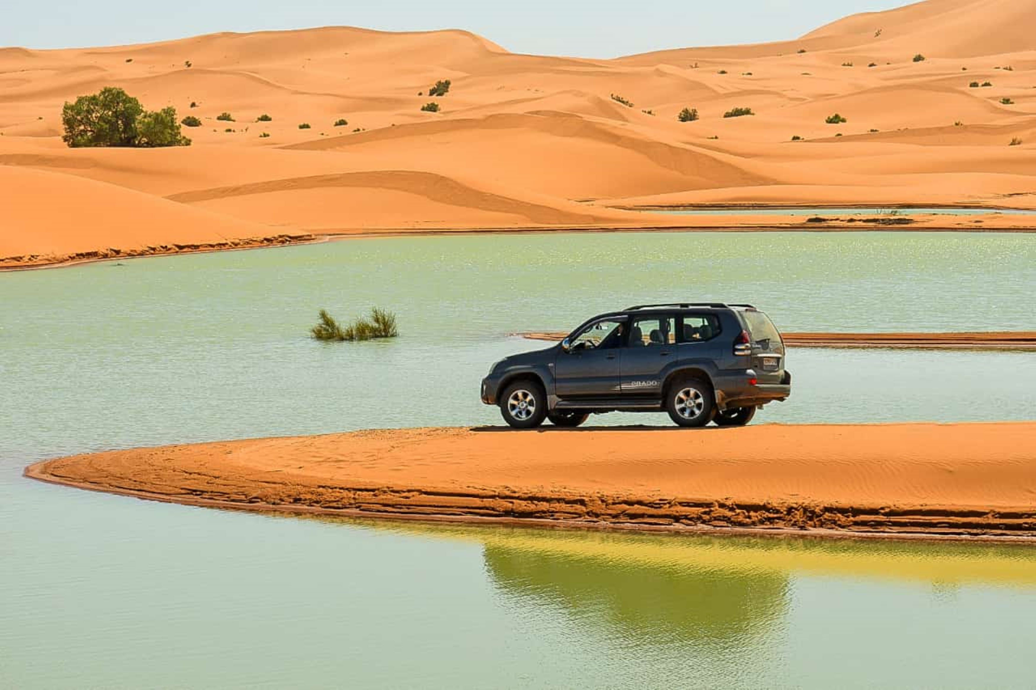 A vehicle parked on a sandy area beside a body of water with sand dunes in the background.