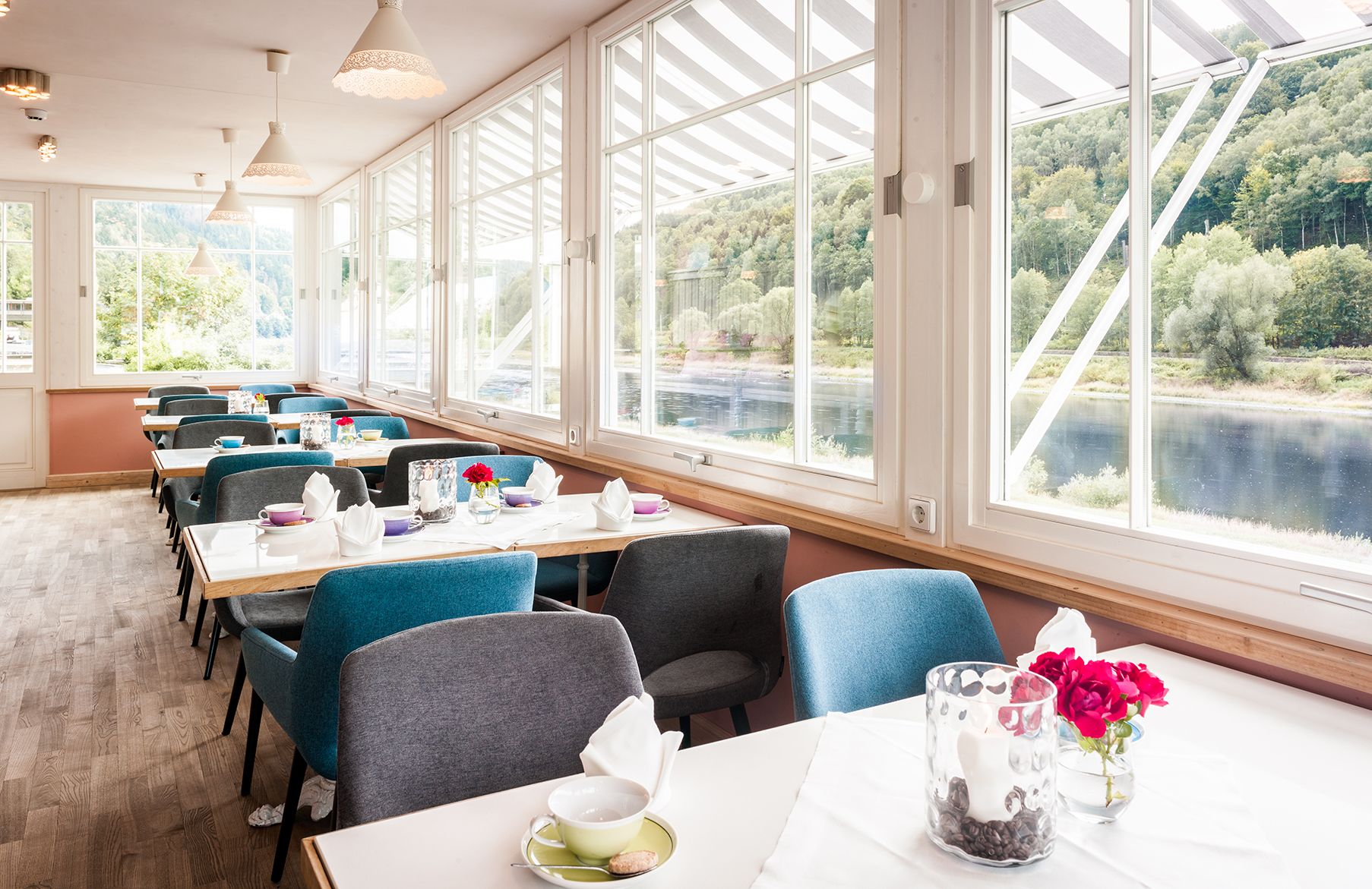 A restaurant with large windows offering a view of a river and forest. Tables are set with chairs and have tableware.