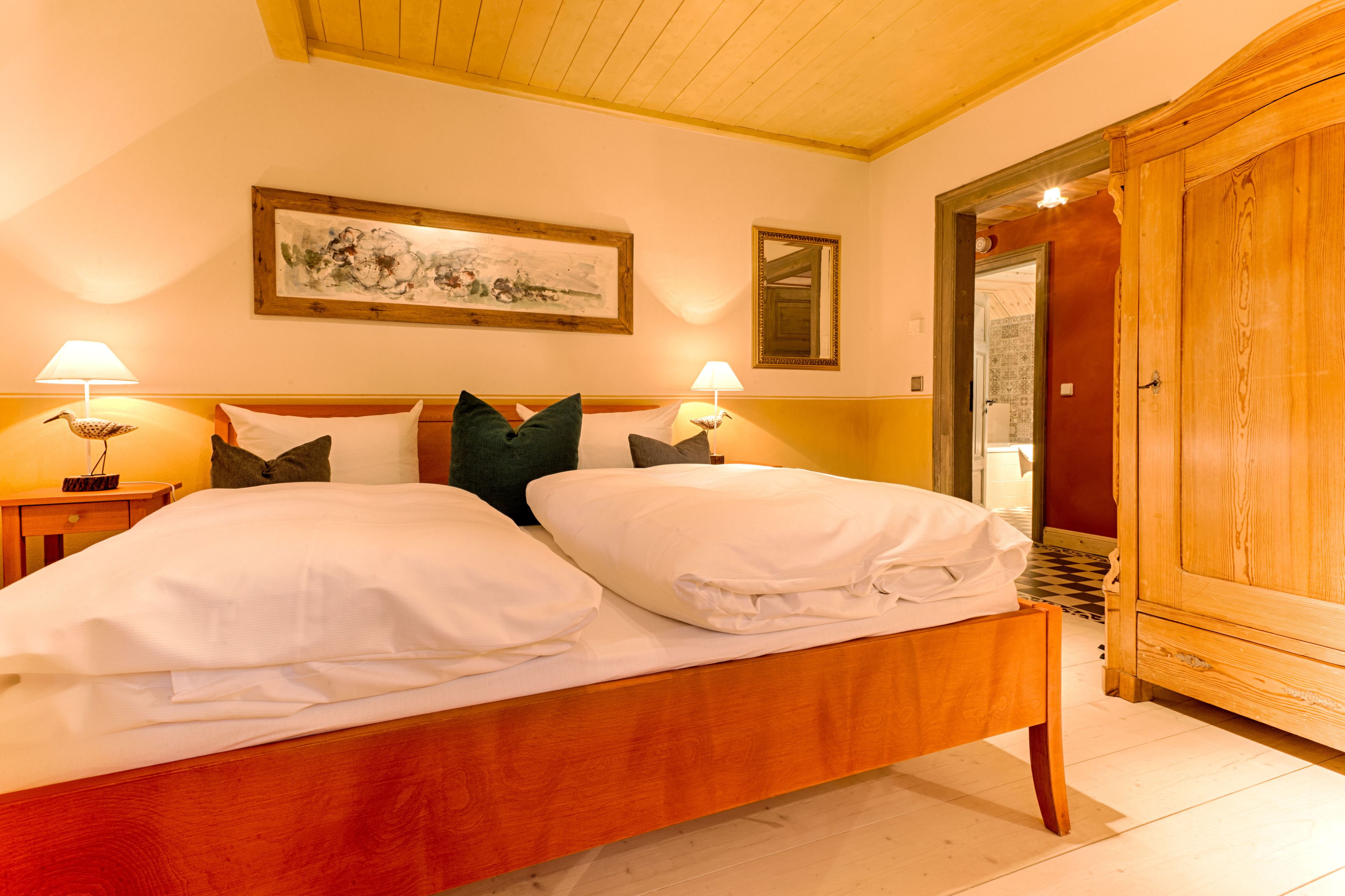 A double bed with white linens, two bedside lamps, a wooden headboard, a framed artwork above the bed, and a wooden wardrobe to the right.