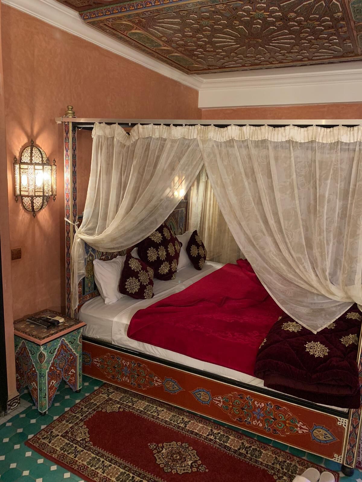 Una cama con dosel adornada con un edredón rojo y dosel blanco, un aplique decorativo y una alfombra estampada.