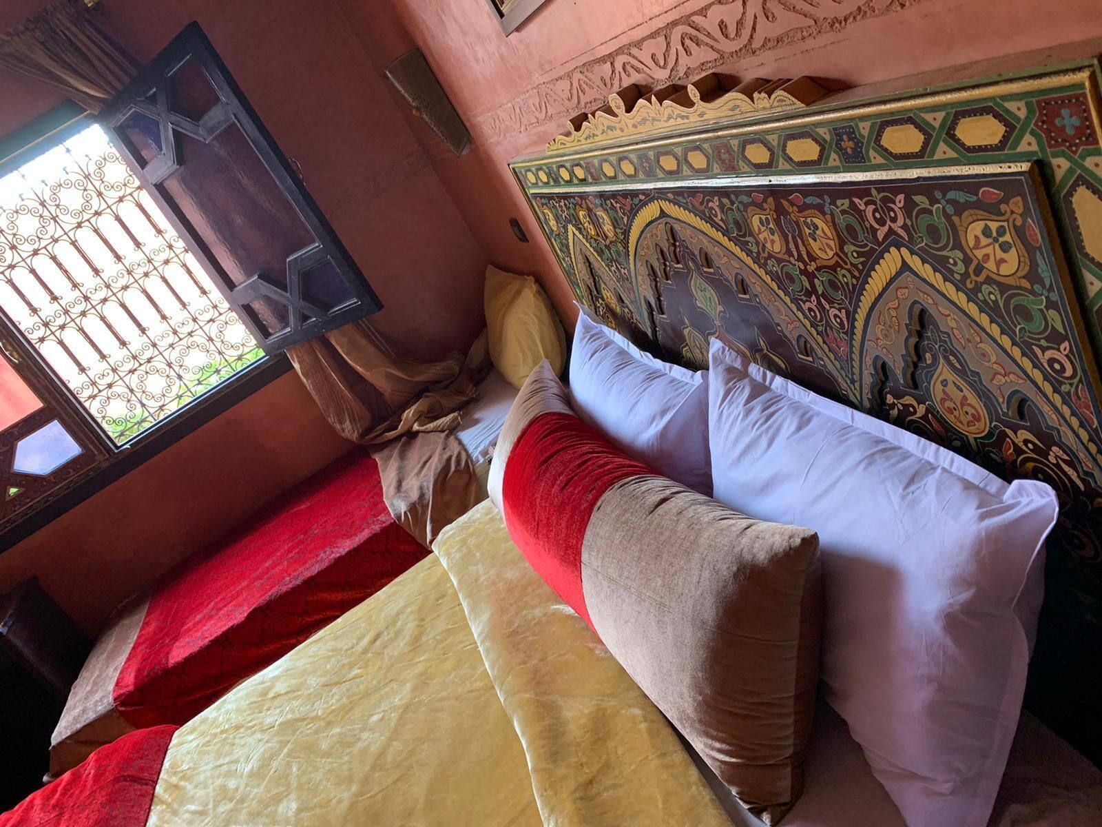 Un lit recouvert d’une couverture rouge et d’une couverture beige, avec deux oreillers, dans une pièce dont la tête de lit est ornée d’un motif et qui possède une fenêtre agrémentée de barreaux en fer décoratifs.