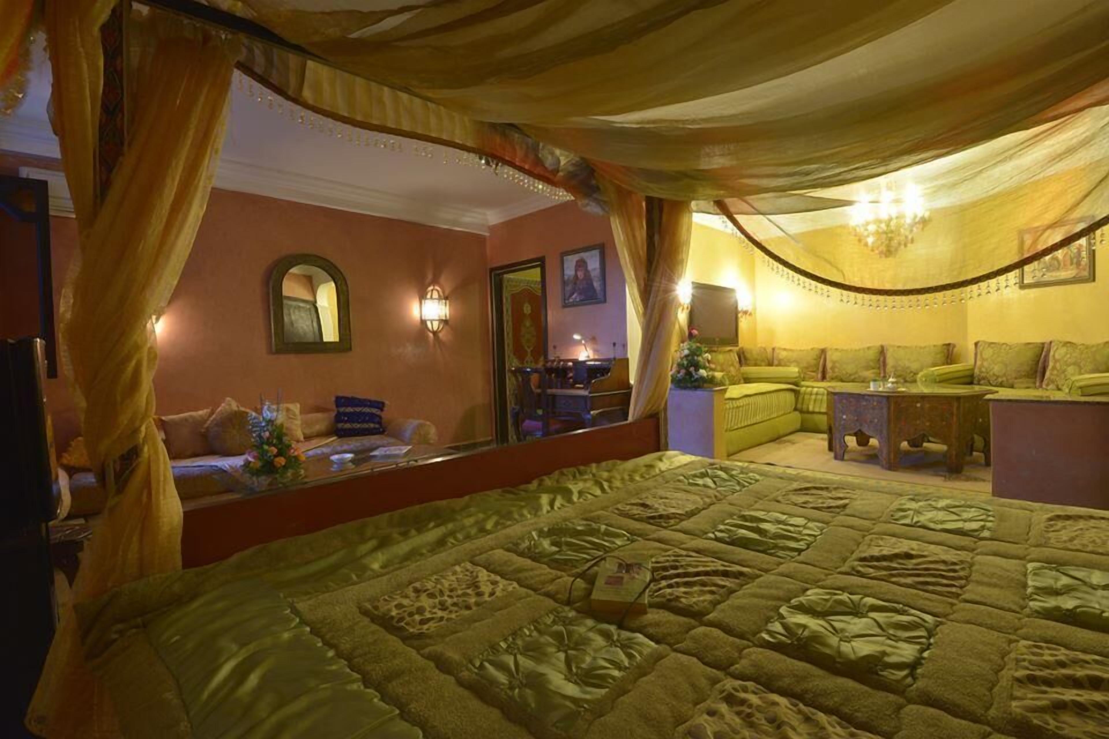 A room with a large bed, a sofa, and a small table. There is a mirror on the wall and a chandelier.