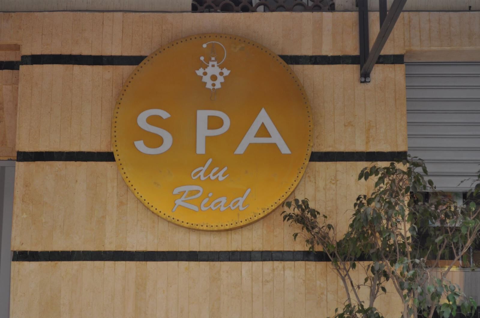 A circular sign with "SPA du Riad" written on it, mounted on a wooden wall with black horizontal stripes.
