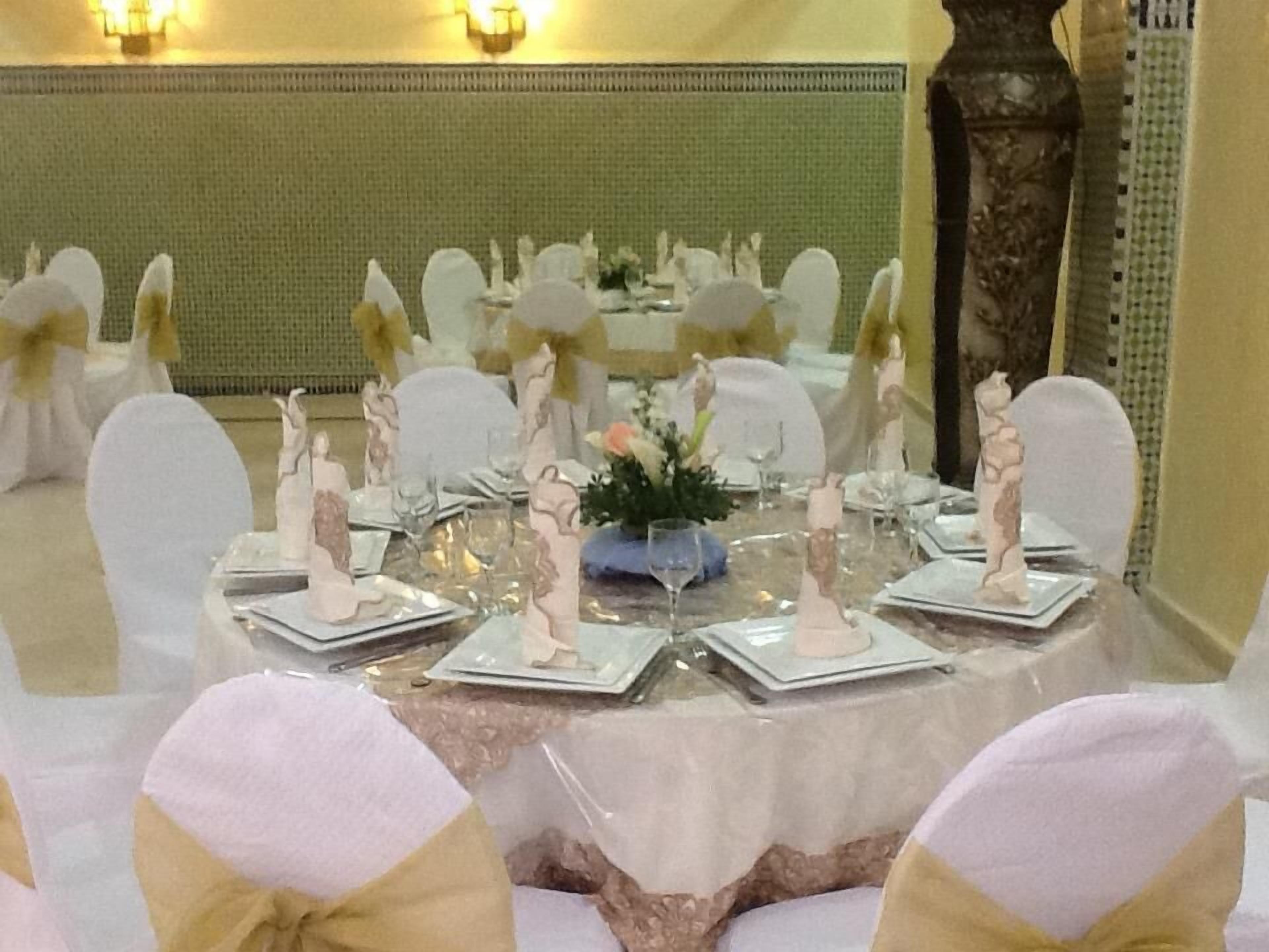Une table de banquet dressée avec de l’argenterie blanche, des verres et des serviettes, entourée de chaises ornées de rubans blancs et dorés.