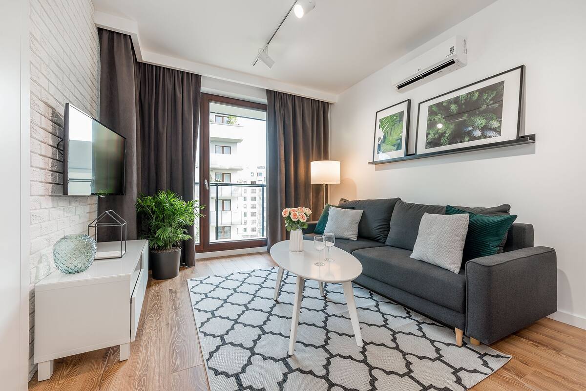 Un salon moderne avec un canapé gris, une table basse blanche, un téléviseur à écran plat et une grande fenêtre avec des rideaux.