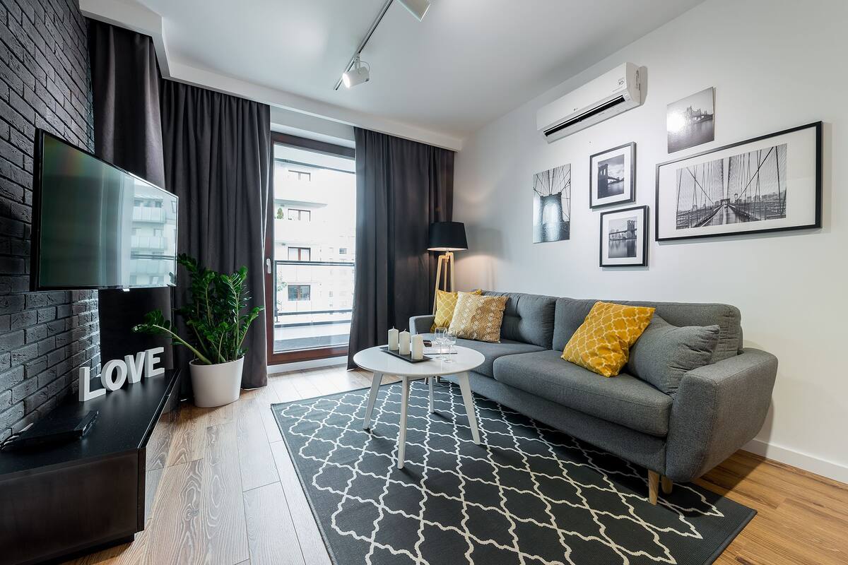 Un salon moderne avec un canapé gris, un tapis à motifs noir et blanc, un téléviseur à écran plat, une plante en pot et un panneau décoratif représentant le mot « AMOUR » accroché au mur.