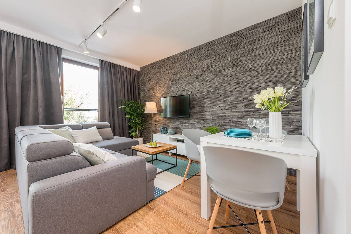 Un salon moderne avec un canapé gris, une table à manger avec des chaises, un téléviseur à écran plat fixé à un mur texturé et une fenêtre avec des rideaux.