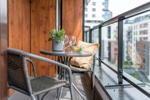 Un balcón con sillas de mimbre, una mesita con una planta en maceta y dos vasos, y vista a edificios a través de una barandilla de vidrio.