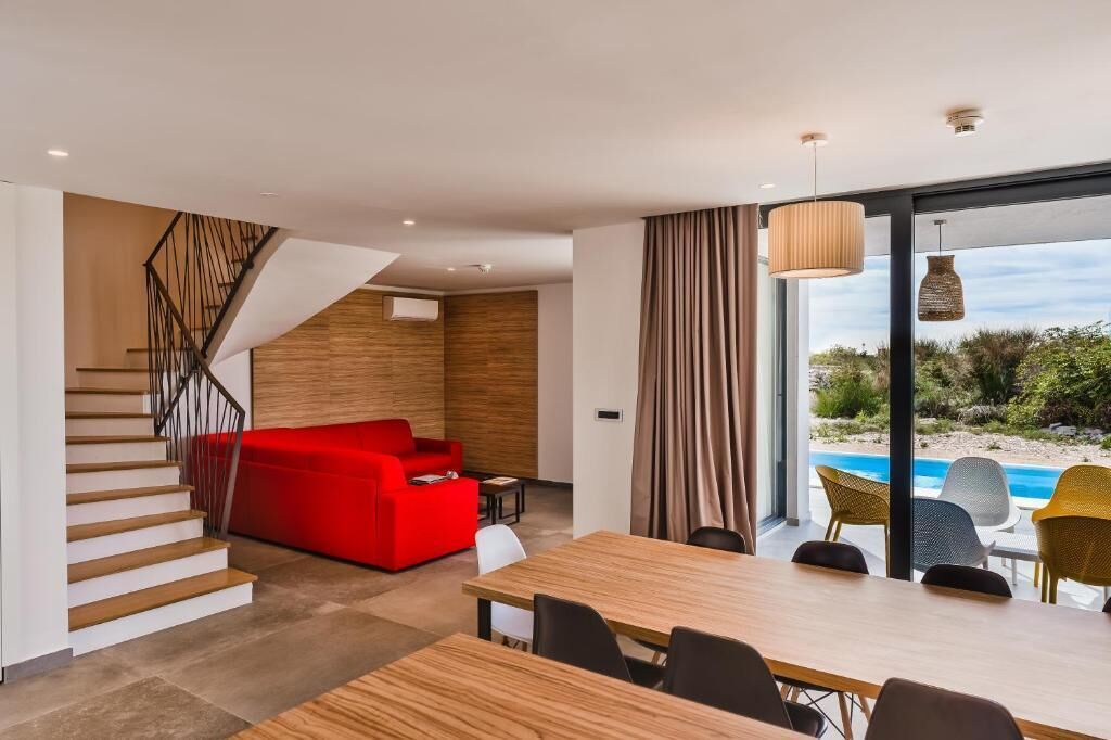 A modern interior with a staircase, a red sofa, a wooden dining table, and a view of a pool.