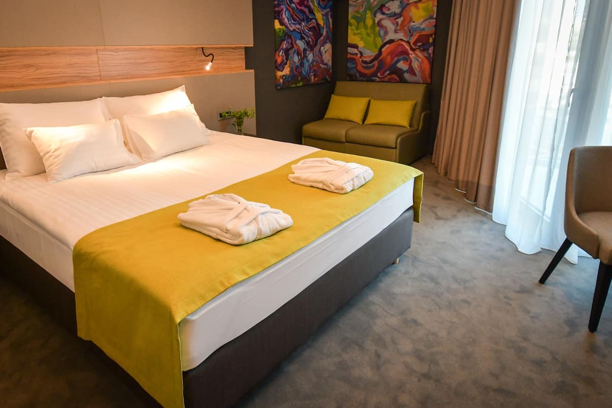 A hotel room with a large bed, two pillows, a yellow bedspread, two towels, a headboard, a painting, a sofa, and a chair.