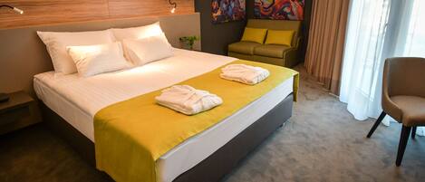 A hotel room with a large bed, two pillows, a yellow bedspread, two towels, a headboard, a painting, a sofa, and a chair.