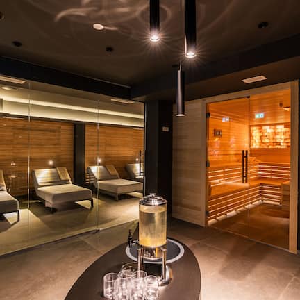 A modern spa room with a glass-enclosed sauna, massage chairs, and a central table with a beverage dispenser.