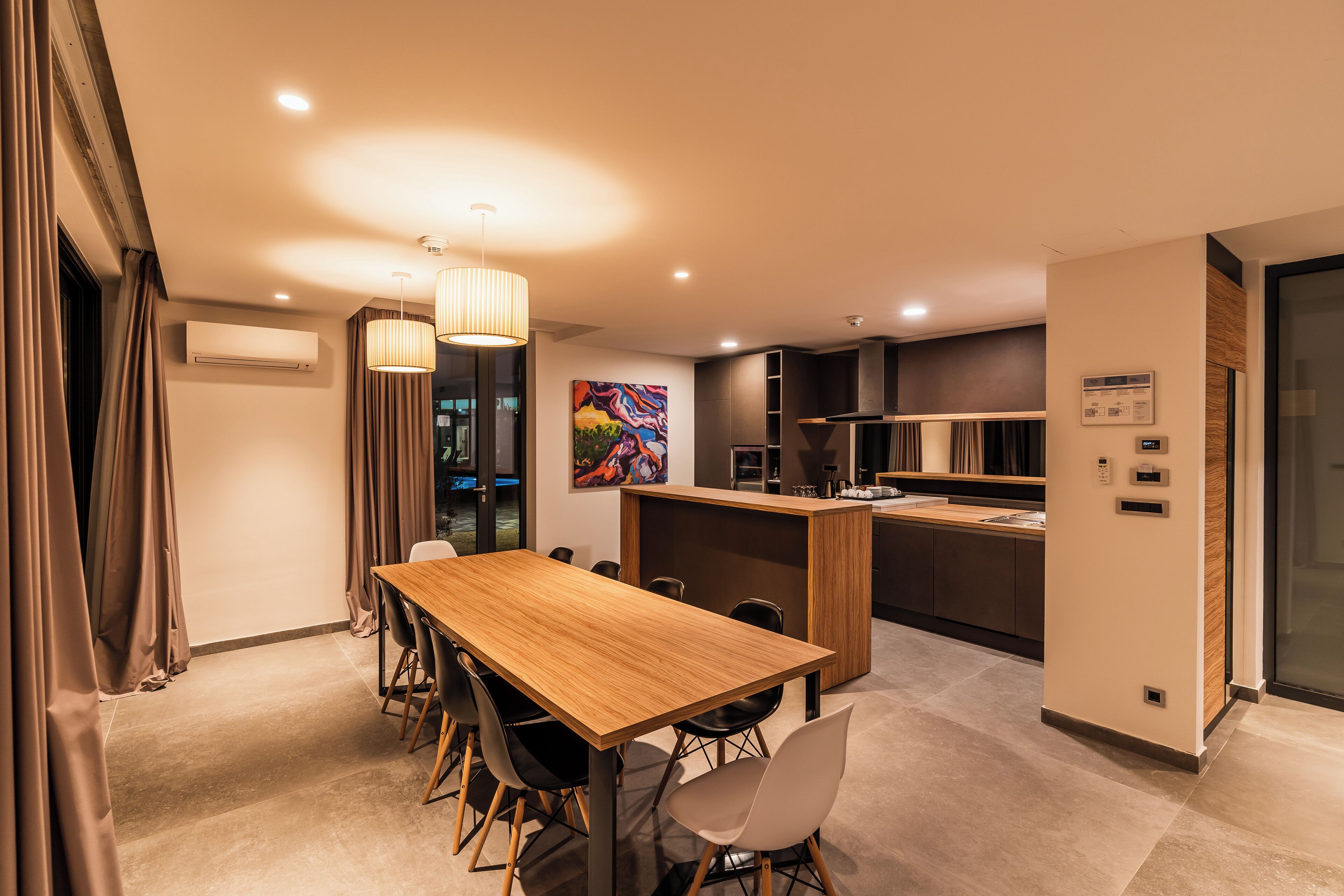 A modern hotel room with a wooden dining table, chairs, a kitchen area, and a large window.