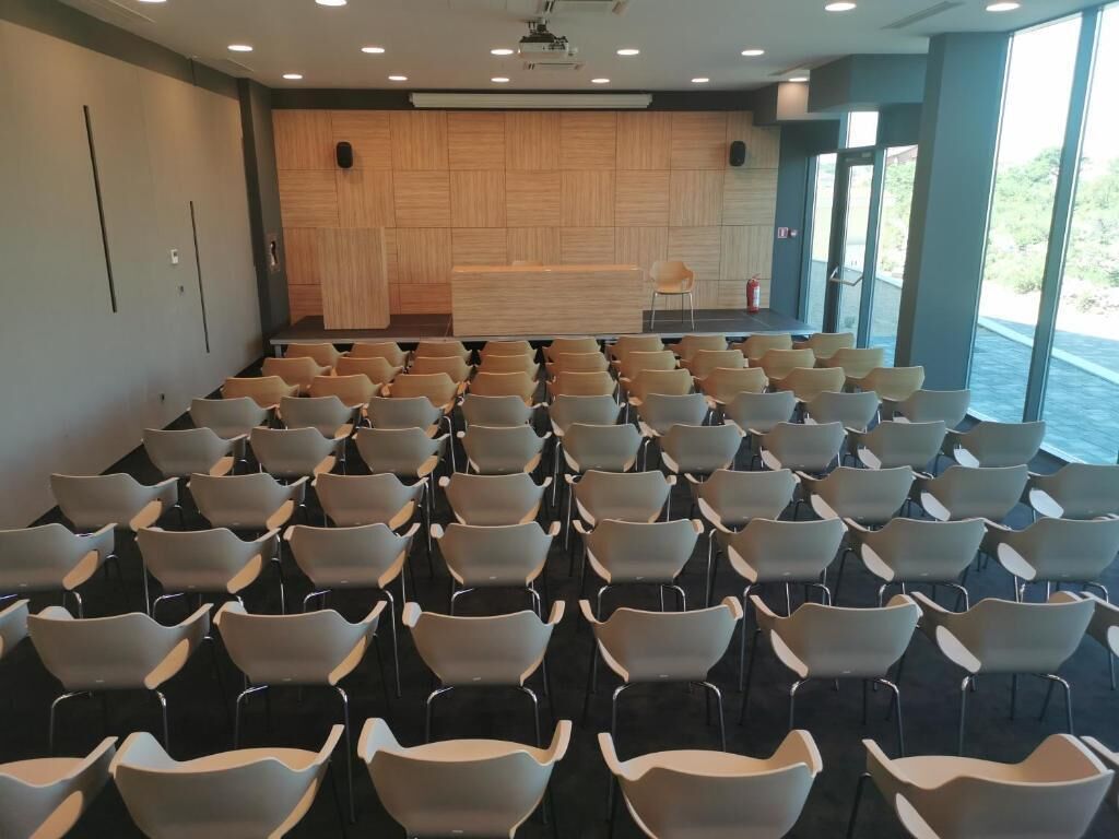 Uma sala de conferências com fileiras de cadeiras, um palco e janelas amplas.