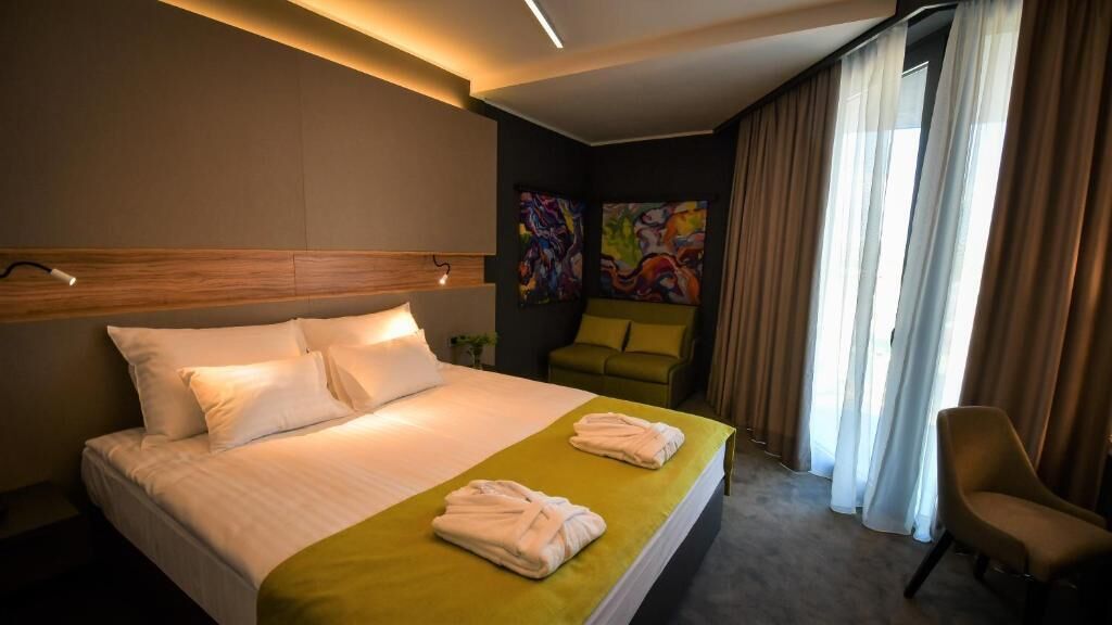 A hotel room with a large bed, a green throw blanket, a chair, and a painting on the wall.