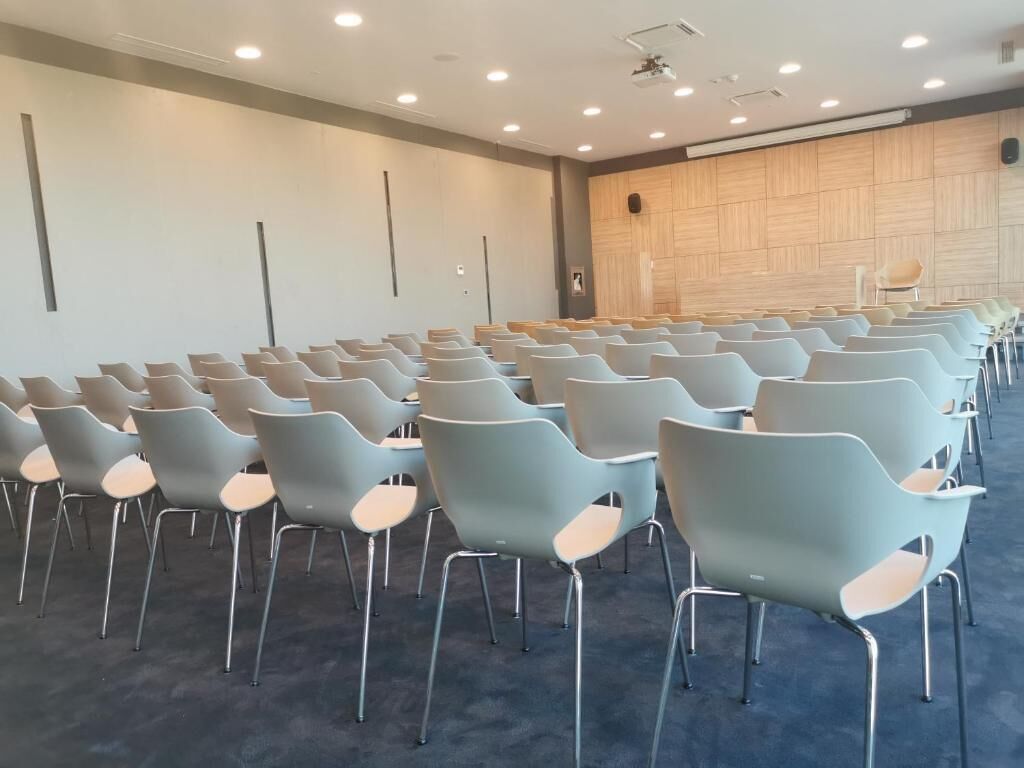 Un salón de conferencias con filas de sillas azul claro y un panel de pared de madera.