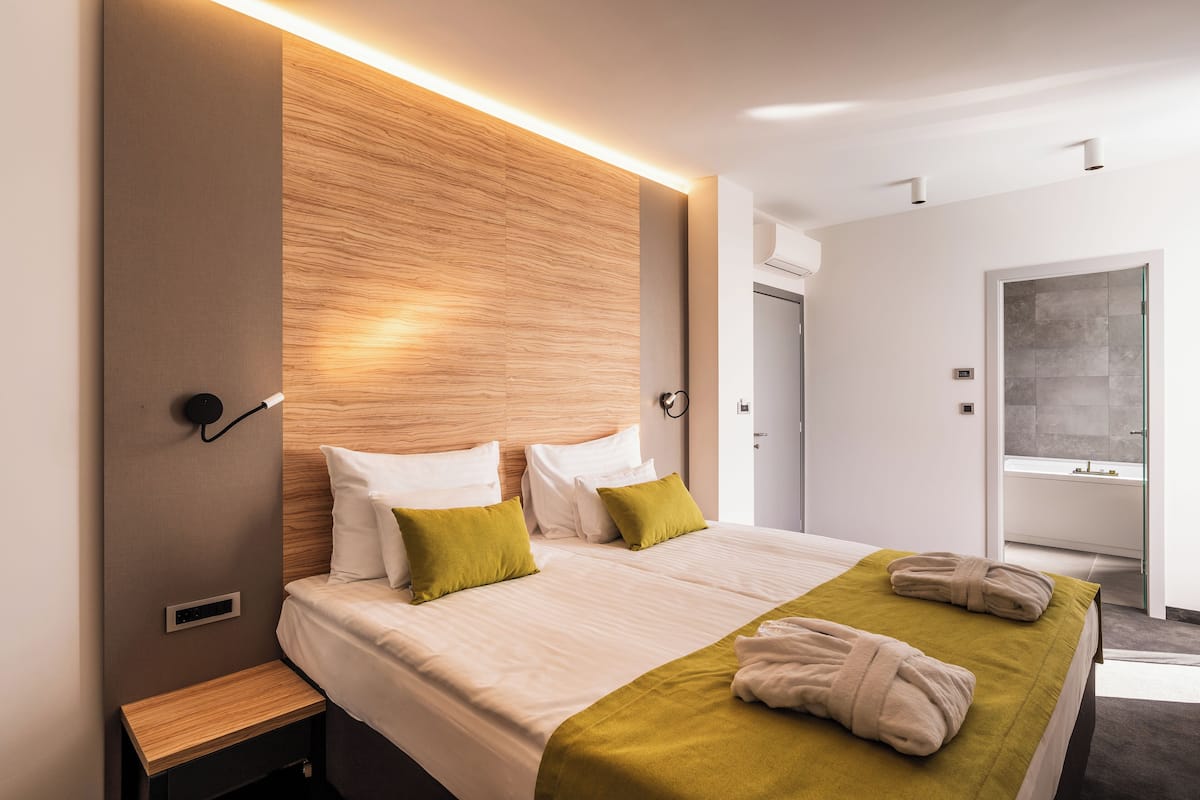 A modern hotel room with a large bed, bedside tables, a wooden headboard, and a bathroom visible through an open door.