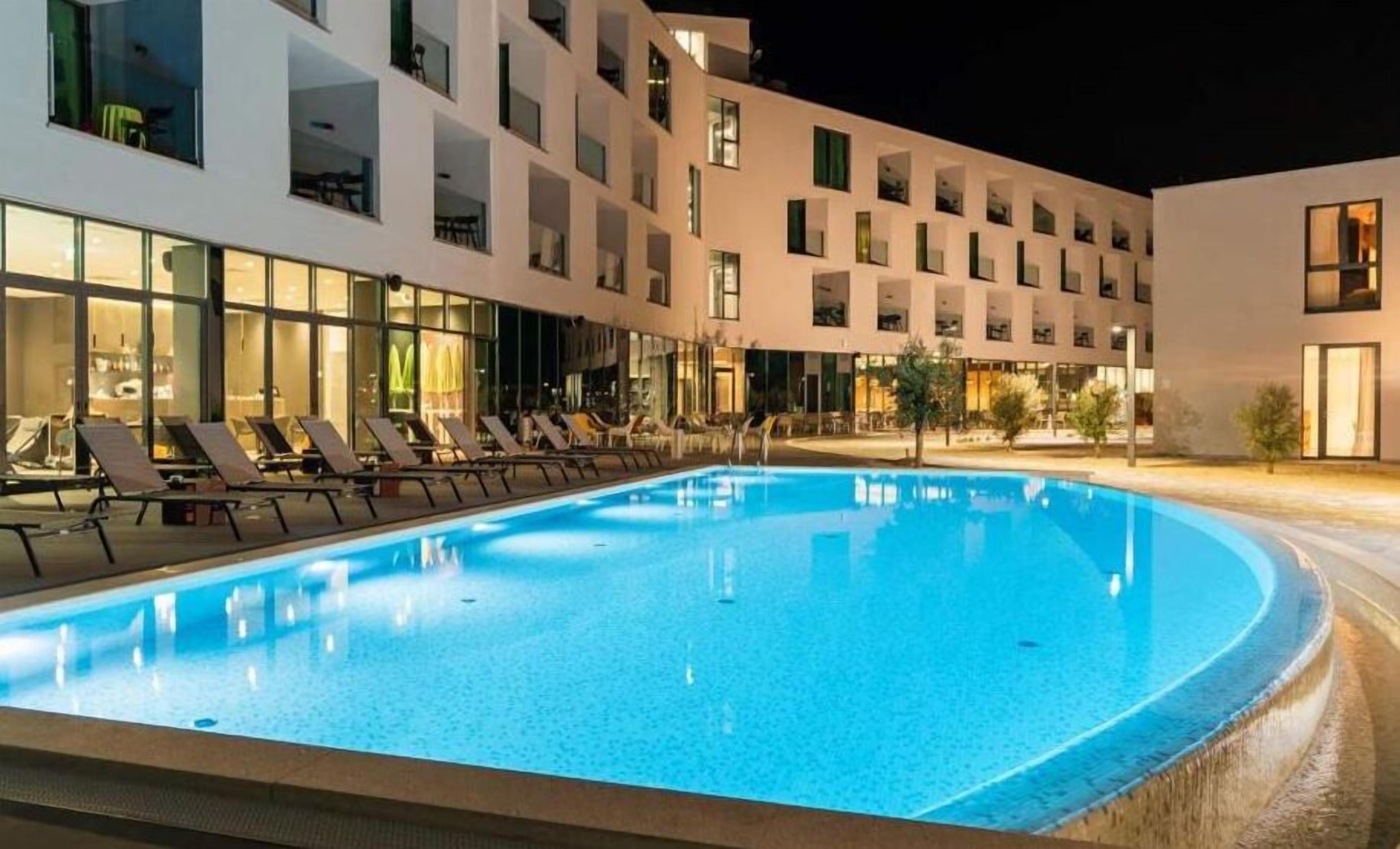 En pool med solstolar och en modern byggnad med flera balkonger i bakgrunden.