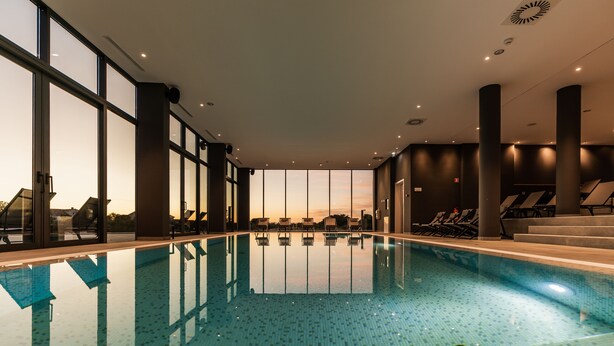 An indoor swimming pool with clear glass walls, a tiled floor, and a sunset view. - Hotel Olea (Novalja)