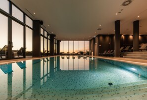 Una piscina interior con paredes de vidrio transparente, suelo de baldosas y vistas al atardecer.