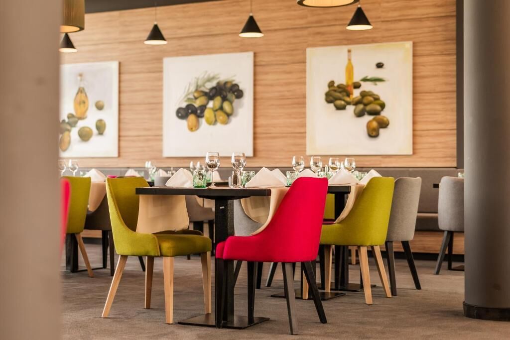 A restaurant with wooden walls, colorful chairs, and framed artwork depicting fruits and olives.