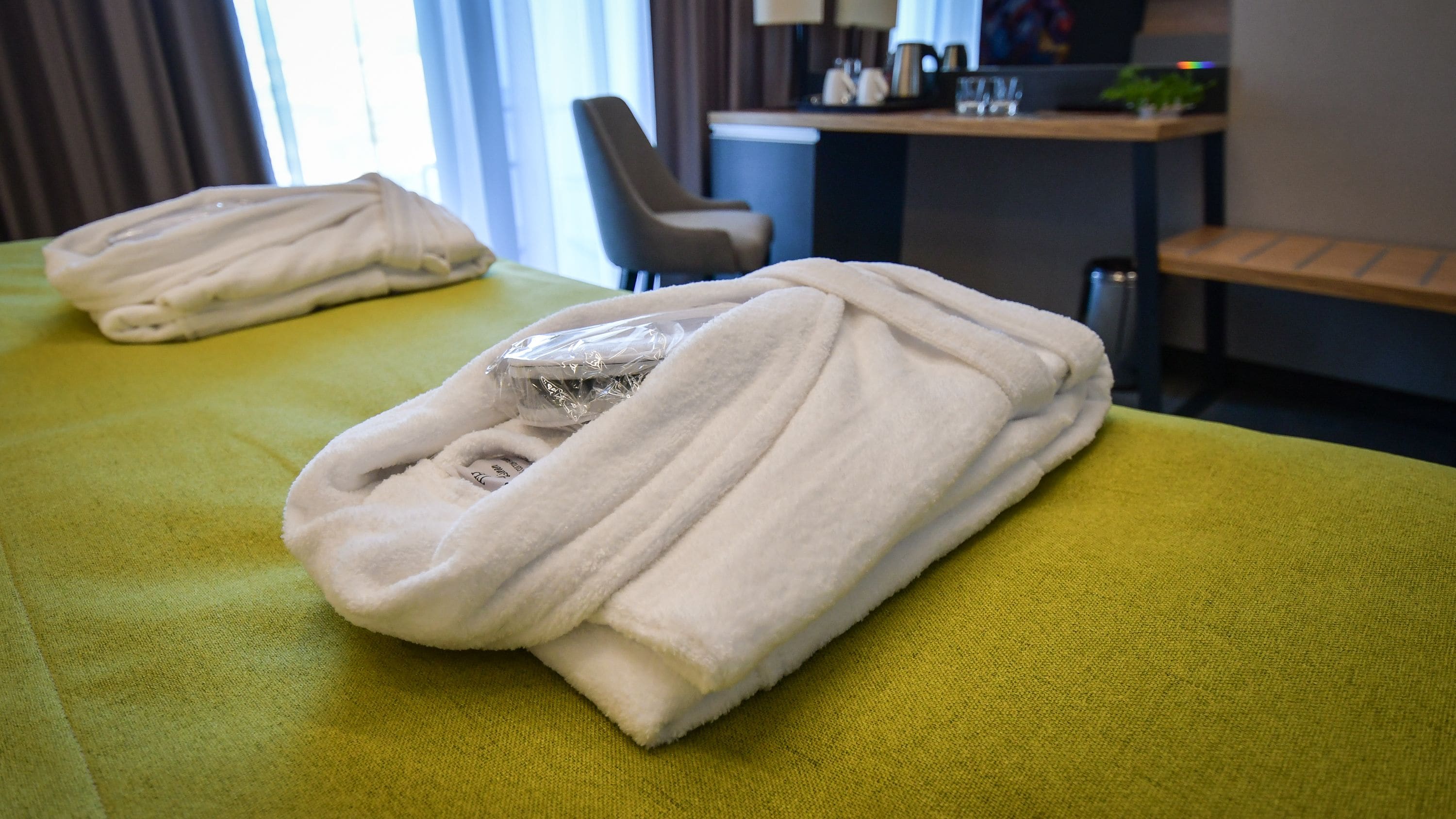 Habitación de hotel con una colcha verde, una toalla blanca doblada y un pequeño objeto envuelto en plástico sobre la cama.