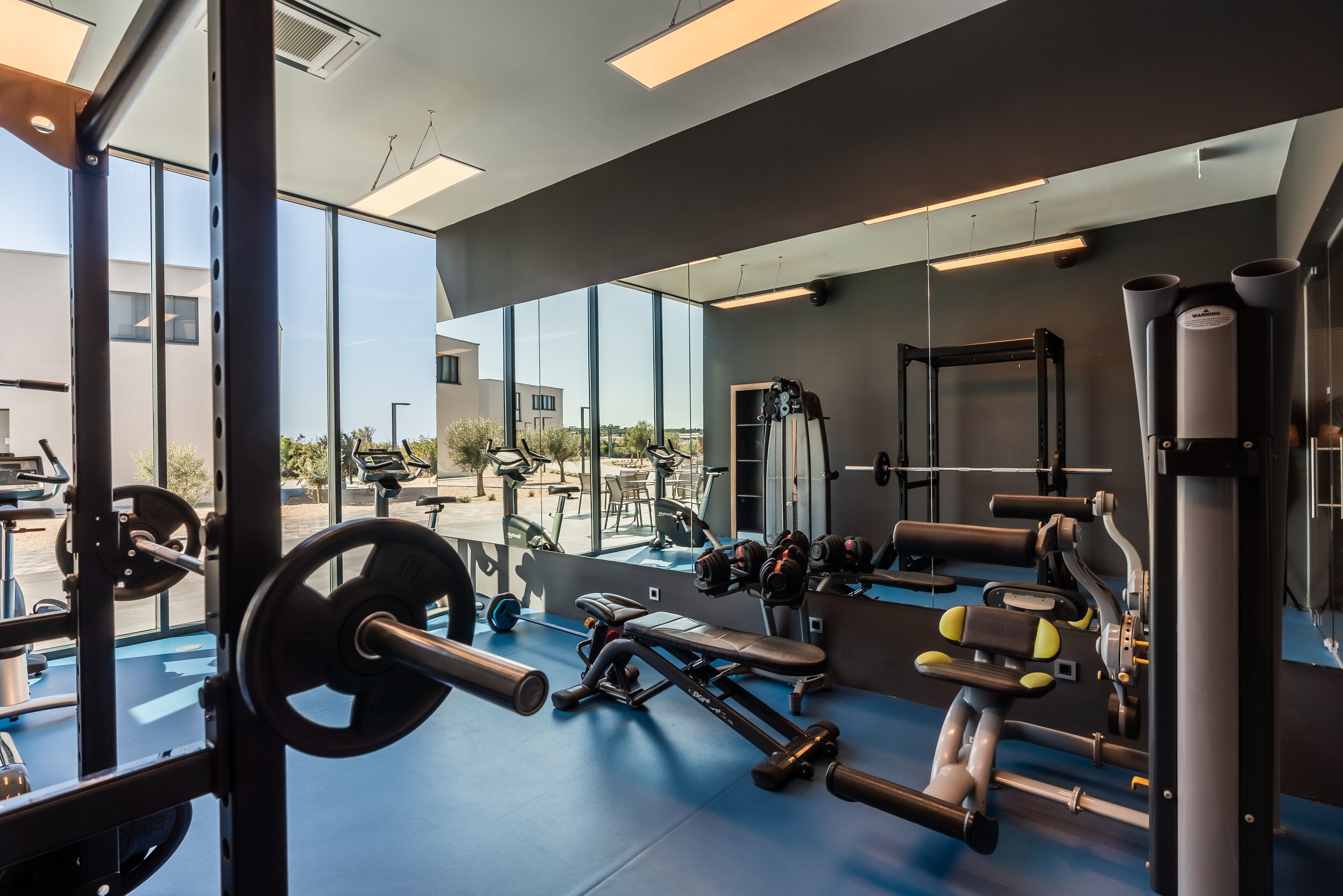 A modern gym with large windows, various exercise equipment, and a blue floor.